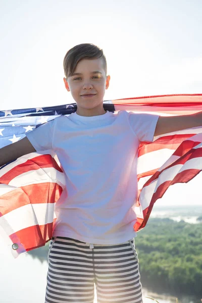 Niño rubio ondeando bandera nacional de EE.UU. al aire libre sobre el cielo azul en la orilla del río —  Fotos de Stock