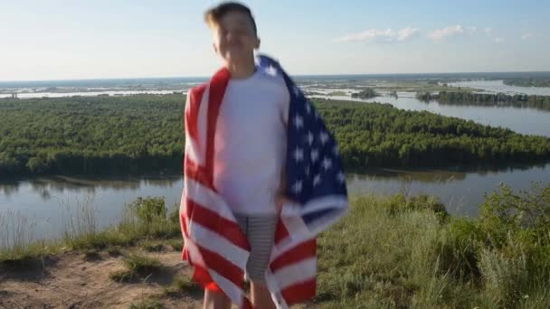 Chlapec mávající národní vlajkou USA venku nad modrou oblohou na břehu řeky — Stock video