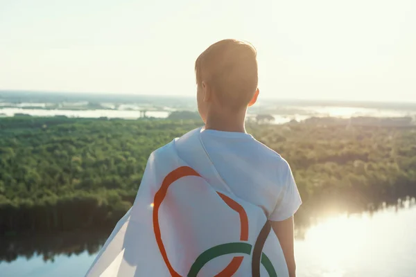 Menino loiro acenando bandeira dos Jogos Olímpicos ao ar livre sobre o céu azul na margem do rio — Fotografia de Stock