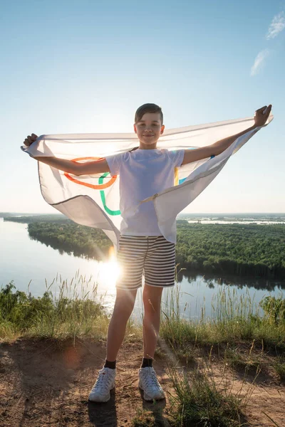 Menino loiro acenando bandeira dos Jogos Olímpicos ao ar livre sobre o céu azul na margem do rio — Fotografia de Stock
