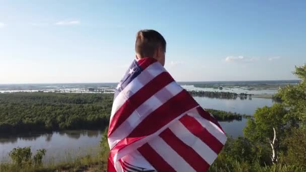 Chlapec mávající národní vlajkou USA venku nad modrou oblohou na břehu řeky — Stock video