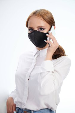 Beyaz stüdyo arka planında izole edilmiş koruyucu maske takan genç kadın akıllı telefonla konuşuyor.