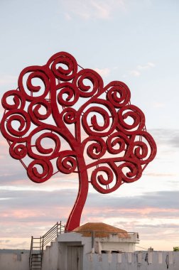 Kıvrımlı dalları olan kırmızı bir ağaç heykeli renkli bir günbatımına karşı siluetlenmiştir..