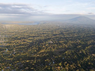 Hava görüntüsü, alacakaranlıkta bulutlu bir gökyüzünün altında büyük bir dağ olan yoğun yeşil tepeleri gösteriyor..