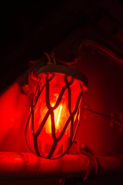 A glowing red alarm light inside an old military submarine. The light is encased in a protective metal cage, creating an intense atmosphere in the confined space