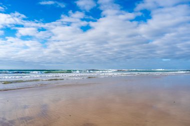 A vast sandy beach stretches along the Great Ocean Road, Australia, with gentle waves lapping the shore under a partly cloudy sky