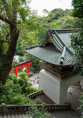 Enoshima-jinja türbesinin çarpıcı kırmızı torii kapısı Enoshima Adası 'nda geleneksel çatılar ve orman tepeleri tarafından çerçevelenen yemyeşil yeşillikler arasında yer alıyor.