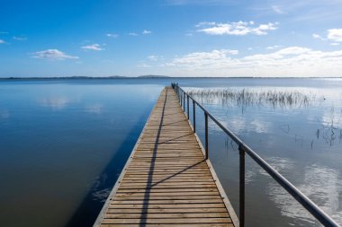 Victoria, Avustralya 'daki Colac Gölü' nün sakin suları üzerinde bir ahşap iskele uzanır. Mavi gökyüzü ve uzak tepeler huzurlu bir atmosfer yaratır.