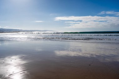 A vast sandy beach stretches along the Great Ocean Road, Australia, with gentle waves lapping the shore under a partly cloudy sky