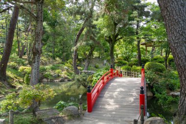 Hiroşima 'daki huzurlu Shukkei-en Japon bahçesinde ağaçlar ve yeşilliklerle çevrili canlı bir kırmızı kemerli köprü bir gölet boyunca uzanır.