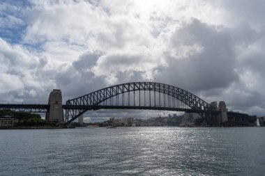 Sydney, Avustralya 'daki Sydney Harbour Köprüsü bulutlu bir gökyüzünün altında liman boyunca uzanır. Çelik kemer köprüsü şehri birbirine bağlıyor, etkileyici yapısının altında yüzen teknelerle gökyüzü ve su manzarası sunuyor.