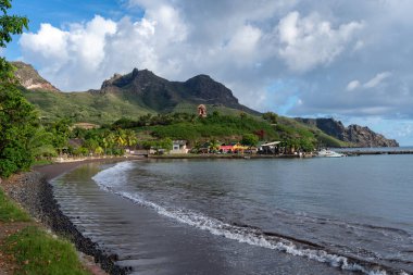 Marquesas Adaları 'nın Nuku Hiva kentindeki Taiohae' nin siyah kumlu plajı tropikal bitki örtüsü, küçük evler ve arka planda dağlar bulunan bir tiki heykeliyle kaplıdır. Fransız Polinezyası
