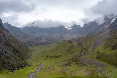 Peru 'daki Humantay Gölü yürüyüşü boyunca uzanan manzaralı bir dağ vadisi engebeli tepeler ve puslu bulutlarla çevrilidir ve çarpıcı bir Andean panoraması yaratır.
