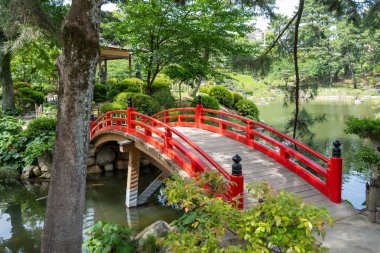 Hiroşima 'daki huzurlu Shukkei-en Japon bahçesinde ağaçlar ve yeşilliklerle çevrili canlı bir kırmızı kemerli köprü bir gölet boyunca uzanır.
