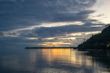Altın bir günbatımı, Fransız Polinezyası 'ndaki Maupiti gölünün sakin sularını dramatik bulutlar ve kıyı şeridi boyunca uzanan siluetli palmiye ağaçlarıyla yansıtır.