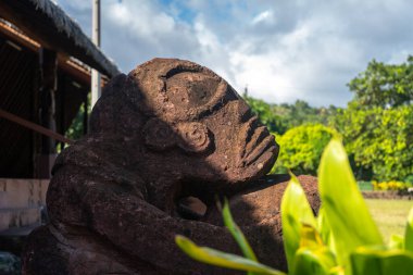 Atuona, Hiva Oa 'da bulunan geleneksel bir Markiz tiki heykeli, Fransız Polinezyası, taştan oyulmuş, yeşil bitkilerle çevrili, dağınık mavi bulutlarla dolu bir gökyüzünün altında.