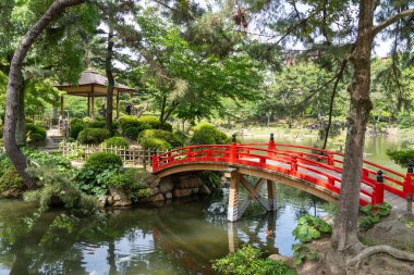 Hiroşima 'daki huzurlu Shukkei-en Japon bahçesinde ağaçlar ve yeşilliklerle çevrili canlı bir kırmızı kemerli köprü bir gölet boyunca uzanır.