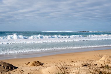 A vast sandy beach stretches along the Great Ocean Road, Australia, with gentle waves lapping the shore under a partly cloudy sky