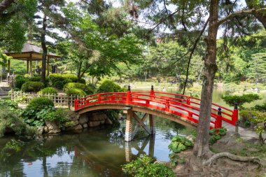 Hiroşima 'daki huzurlu Shukkei-en Japon bahçesinde ağaçlar ve yeşilliklerle çevrili canlı bir kırmızı kemerli köprü bir gölet boyunca uzanır.
