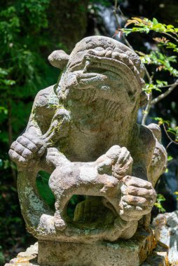 Hiroşima 'daki Mitaki-Dera Tapınağı' nda ağaçlar ve ahşap film plaklarıyla çevrili yıpranmış bir Komanu koruyucu heykeli gün ışığında dinleniyor.