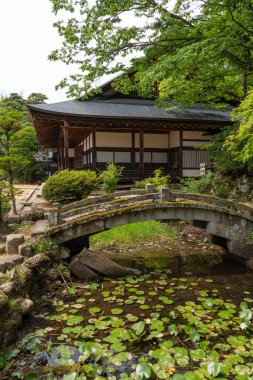 Japonya 'nın Takayama kentindeki Zenno-ji Tapınağı' nın barışçıl bir manzarası. Çerçevesi gür ağaçlar, geleneksel bir taş köprü ve yüzen zambaklarla dolu bir gölet.