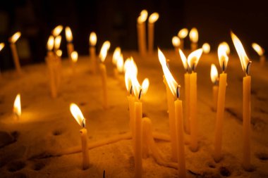 Bir grup yanan mum kumdan bir yatakta duruyor, titreyen alevleri sıcak, altın bir parıltı saçıyor. Balmumu, ateşin ışığı yansımayı ve bağlılığı sembolize eden barışçıl ve ruhani bir atmosfer yaratırken damlıyor.