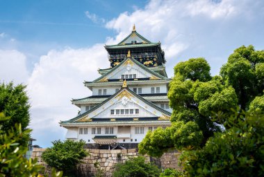 Osaka Kalesi 'nin görkemli ana kulesi parlak bir gökyüzü altında yeşil ağaçların üzerinde yükseliyor. Geleneksel Japon mimarisi ve altın süslemeleri sergiliyor.