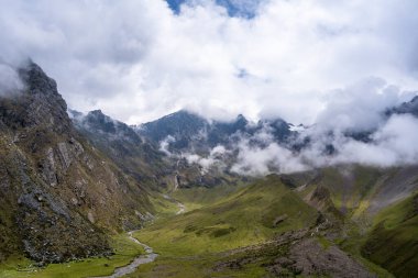 Peru 'daki Humantay Gölü yürüyüşü boyunca uzanan manzaralı bir dağ vadisi engebeli tepeler ve puslu bulutlarla çevrilidir ve çarpıcı bir Andean panoraması yaratır.