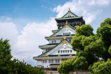 Osaka Kalesi 'nin görkemli ana kulesi parlak bir gökyüzü altında yeşil ağaçların üzerinde yükseliyor. Geleneksel Japon mimarisi ve altın süslemeleri sergiliyor.
