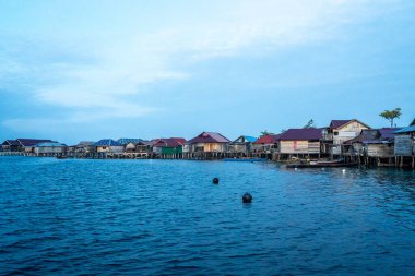 Pulau Papan 'ın ahşap balıkçı evleri, Endonezya, Malenge Adası yakınlarındaki sakin deniz sularıyla çevrili yumuşak günbatımı ışığı altında parlıyor.