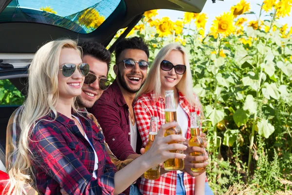 Amici che bevono birra tostatura clink bottiglie seduti in auto tronco campagna esterna — Foto Stock