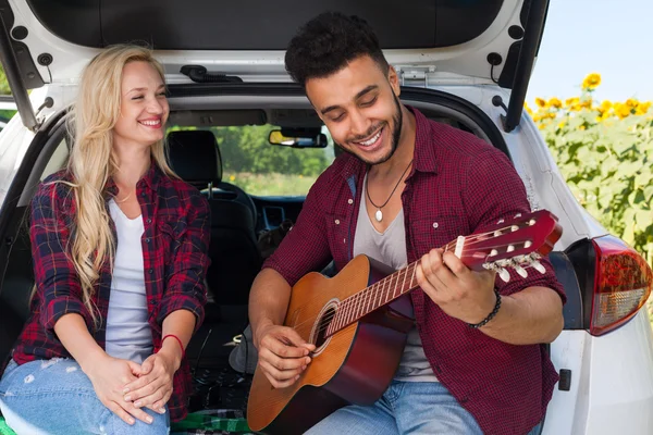 Young girl listening boyfriend playing guitar couple sitting car trunk outdoor countryside — Stock fotografie