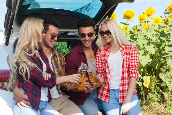 Amici che bevono birra tostatura clink bottiglie seduti in auto tronco campagna esterna — Foto Stock