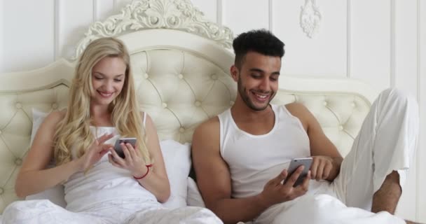 Pareja acostado cama usando el teléfono inteligente, mezclar raza hombre mujer sonrisa mañana dormitorio — Vídeo de stock