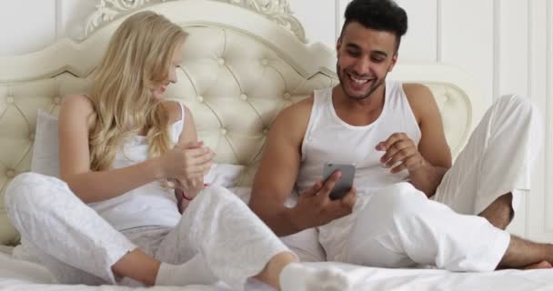 Couple couché lit en utilisant le téléphone intelligent, mélanger course homme femme sourire matin chambre — Video