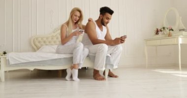 Çift akıllı telefon oturma yatak karışımı yarış adam kadın kucaklamak gülümseme sabah yatak odası kullanarak