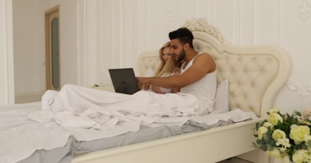 Pareja acostado cama usando ordenador portátil, mezclar raza hombre mujer sonrisa mañana dormitorio — Vídeo de stock