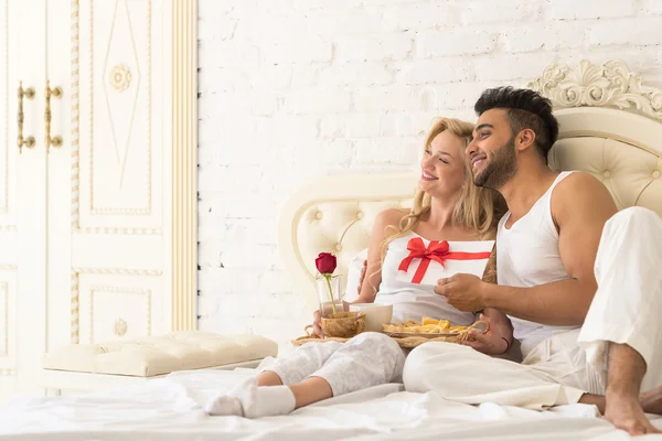 Jeune couple heureux allongé au lit, l'homme hispanique donne à la femme surprise présente enveloppe avec ruban — Photo