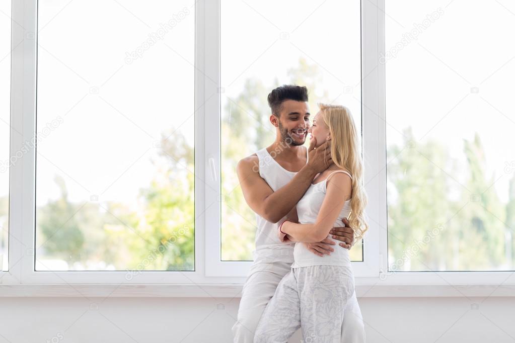 Young Beautiful Couple Stand Near Big Window Embrace Kissing, Happy Hispanic Man And Woman