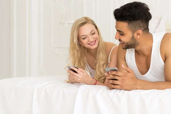Junges Paar liegt im Bett, glückliches Lächeln spanischer Mann und Frau mit Handy — Stockfoto