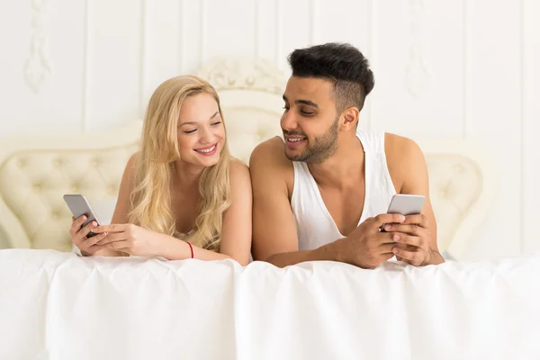 Junges Paar liegt im Bett, glückliches Lächeln spanischer Mann und Frau mit Handy — Stockfoto