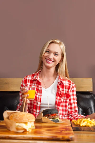 Hermosa rubia mujer sentado en café celebrar naranja jugo orden carne hamburguesa servida en papel en madera mesa — Foto de Stock