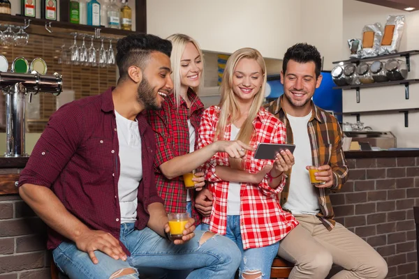 Amigos de la gente usando el teléfono inteligente celular, bebiendo jugo de naranja hablando riéndose sentado en el mostrador del bar, hombre y mujer de raza mixta —  Fotos de Stock
