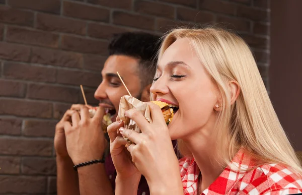 Hombre joven y mujer comiendo hamburguesas de comida rápida sentados en la mesa de madera en el café — Foto de Stock