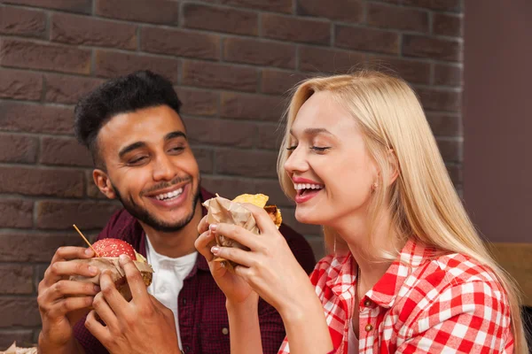 Hombre joven y mujer comiendo hamburguesas de comida rápida sentados en la mesa de madera en el café — Foto de Stock