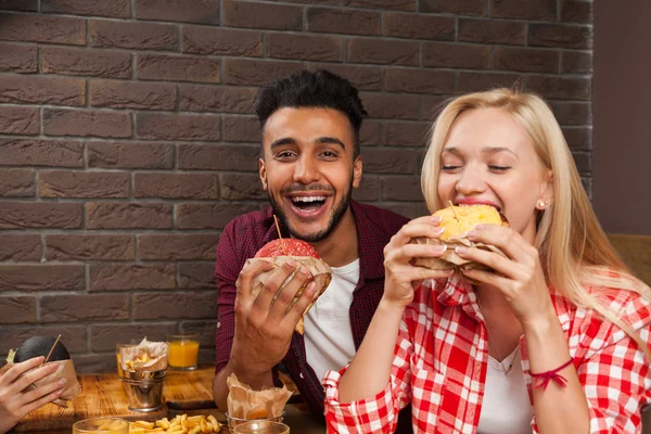 Hombre joven y mujer comiendo hamburguesas de comida rápida sentados en la mesa de madera en el café — Foto de Stock