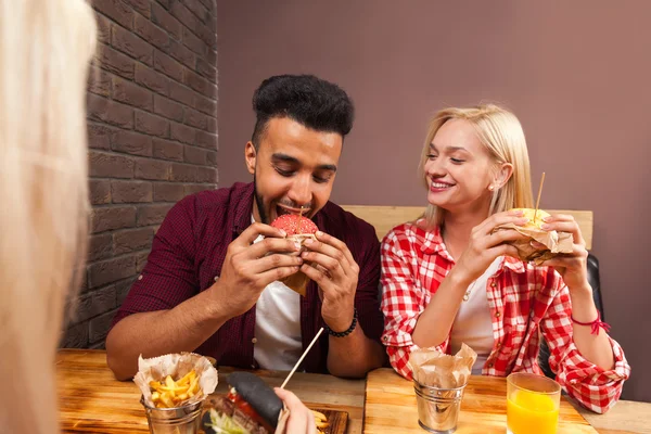 Молодой человек и женщина едят гамбургеры быстрого питания, сидя за деревянным столом в кафе — стоковое фото
