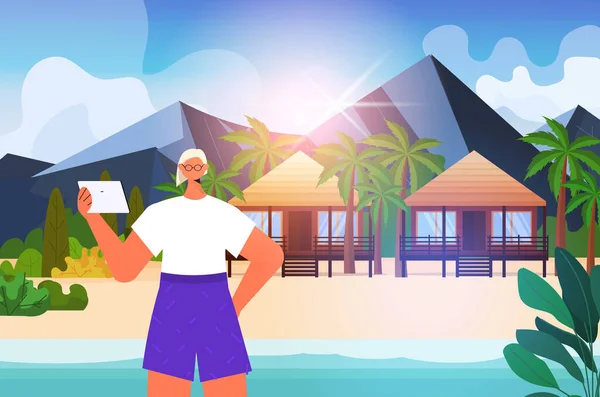 Seniorka relaksująca się na tropikalnej plaży starsza pani korzystająca z tabletu wakacje aktywna starość koncepcja — Wektor stockowy