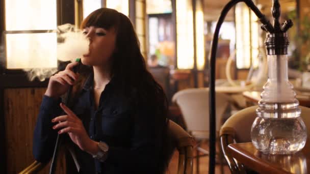 Όμορφη νεαρή γυναίκα εισπνεόμενου ναργιλέ. κορίτσι κάπνισμα ναργιλέ στο καφέ. Θερμό φως — Αρχείο Βίντεο