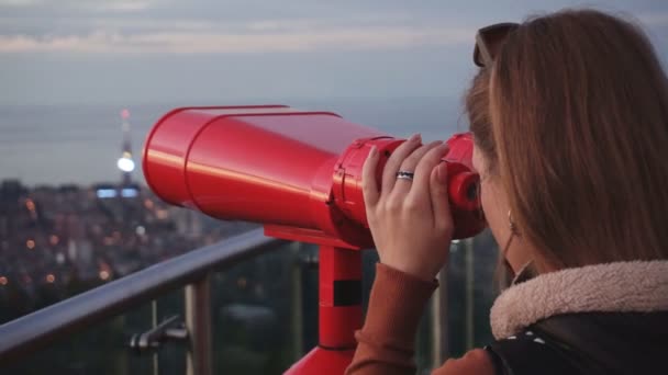 Cestování: mladá žena, která se při západu slunce dívá na město pomocí dalekohledu ovládajícího mince. Snímek zblízka, kapesní, pomalý pohyb 60fps — Stock video